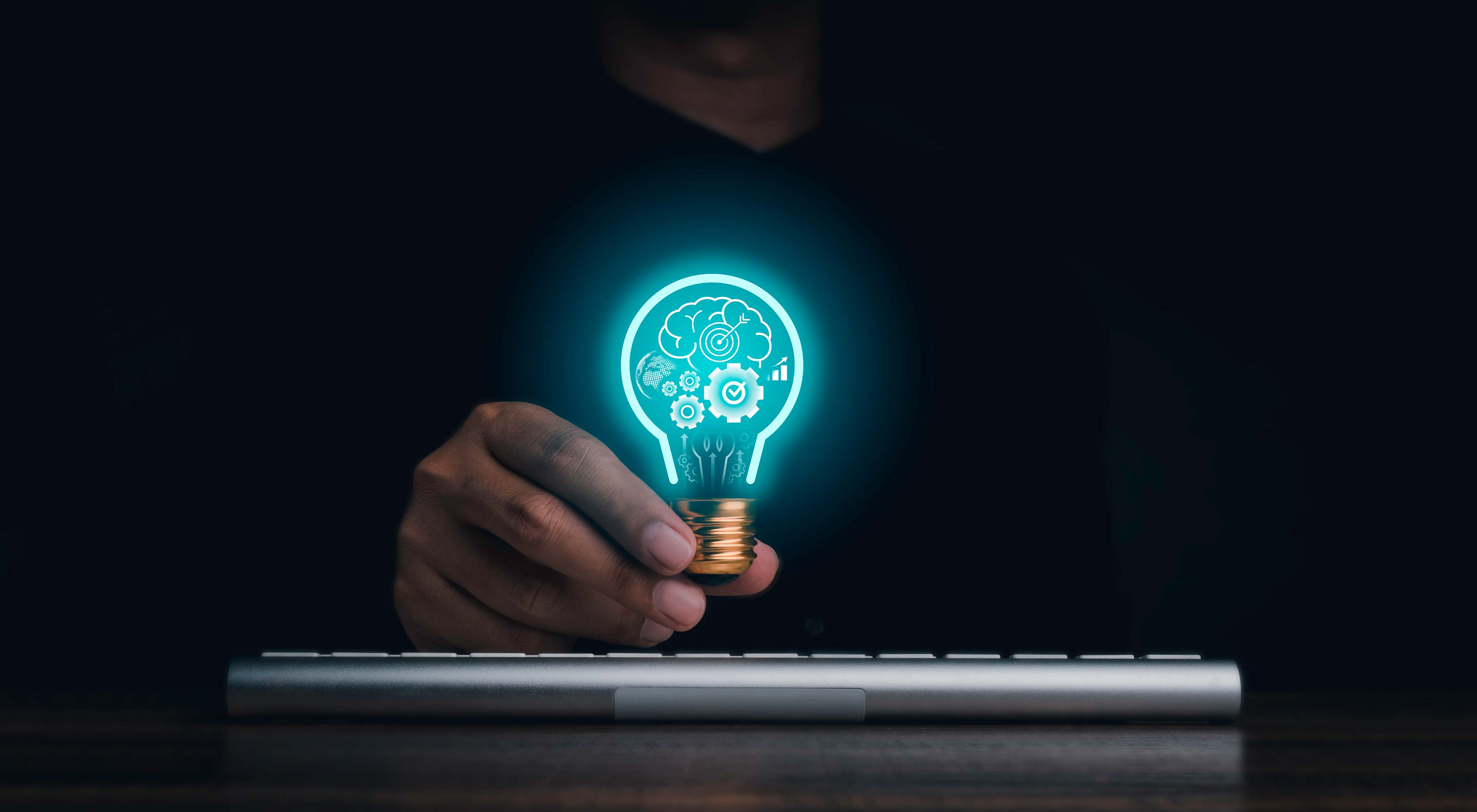 Photo of a man holding a light bulb over a keyboard, with graphics inside the light bulb such as gears and a brain