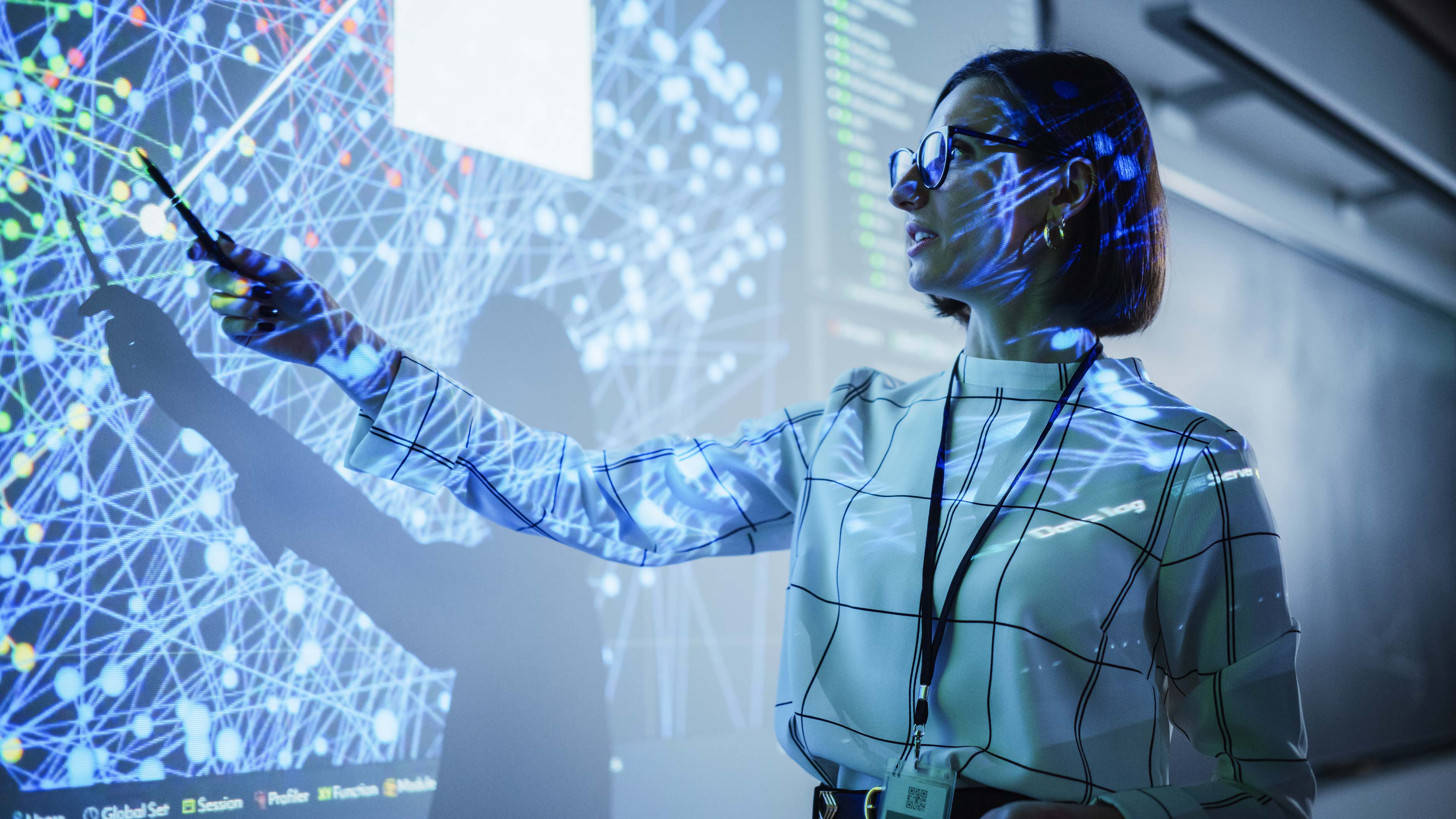 A woman in glasses points to a large screen displaying a complex network of interconnected nodes, illuminated by blue and white light patterns.
