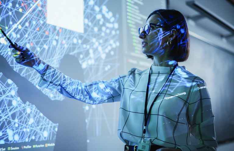 A woman in glasses points to a large screen displaying a complex network of interconnected nodes, illuminated by blue and white light patterns.
