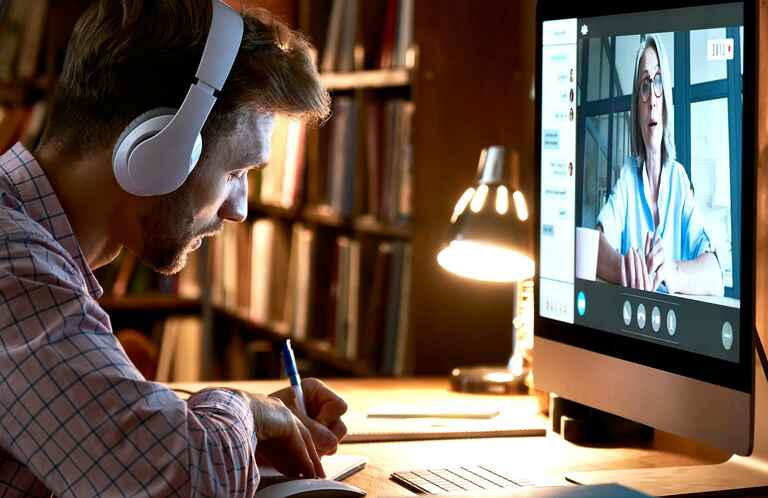A man wearing headphones takes notes while watching a woman speaking on a computer screen in a virtual meeting, with a bookshelf and lamp in the background.