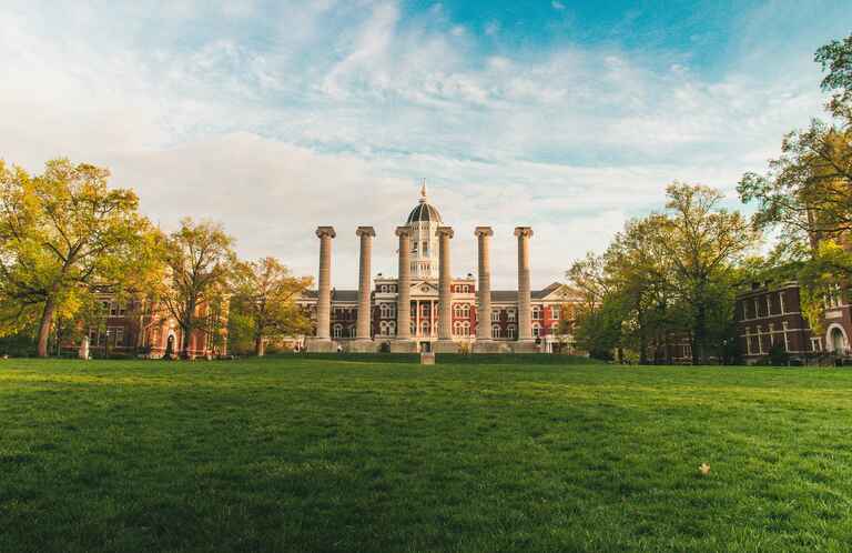 A grassy quad with six stone columns in the center, flanked by trees and historic red-brick buildings, with a domed building in the background under a blue sky.