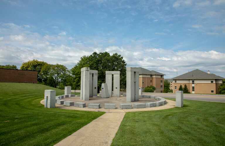 A stone circle monument with tall rectangular pillars and shorter blocks arranged in a circular formation on a grassy lawn, with buildings and trees in the background.