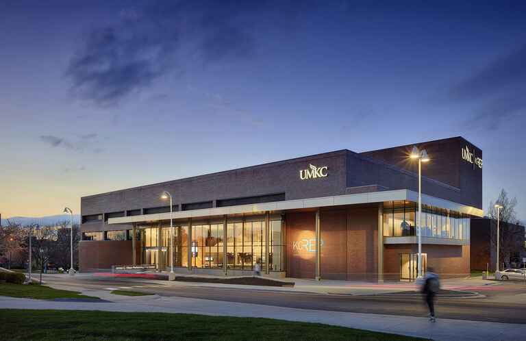 The James C. Olson Performing Arts Center at UMKC, a modern building with large glass windows and brick walls, illuminated at dusk with a person walking outside.