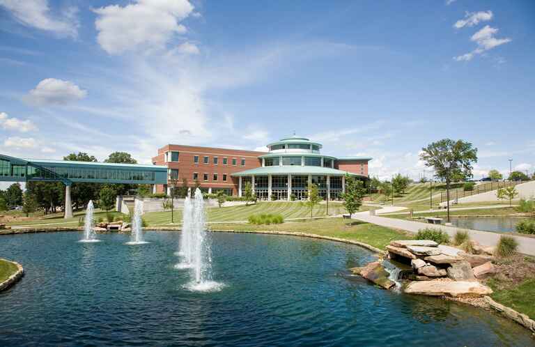 A scenic view of the UMSL campus featuring a modern brick building with large windows, a pond with fountains, and a pedestrian bridge under a clear blue sky.