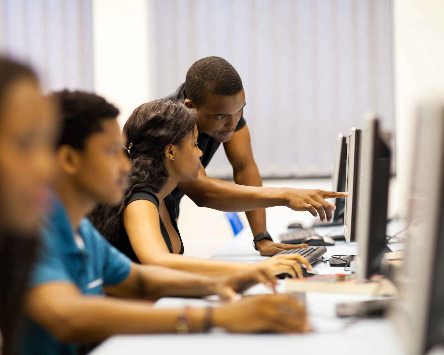 an instructor points to a computer screen in a computer lab with a class full of students