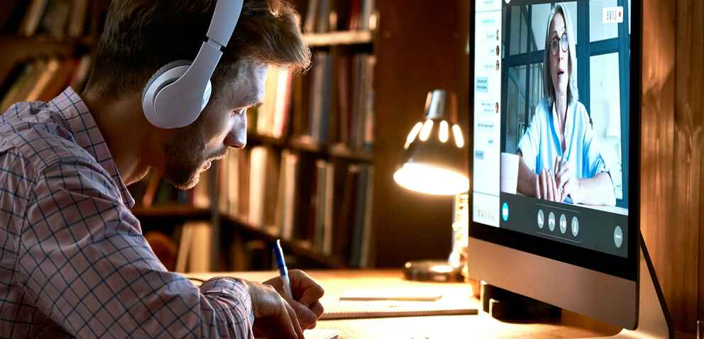A man wearing headphones takes notes while watching a woman speaking on a computer screen in a virtual meeting, with a bookshelf and lamp in the background.